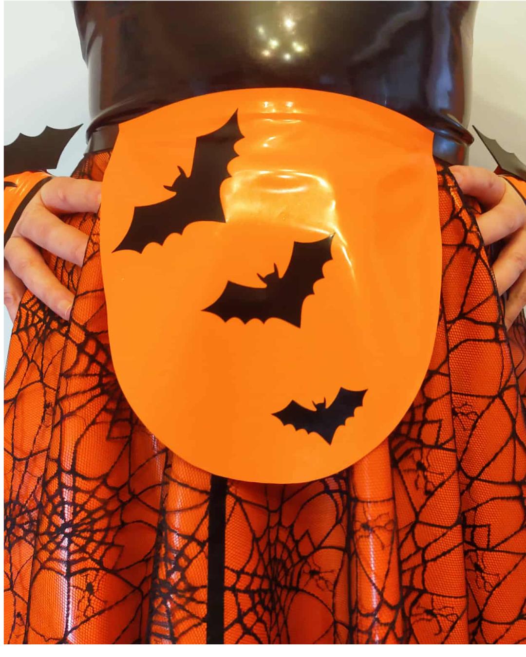 An orange latex apron featuring three black bat silhouettes, worn over an orange and black spiderweb-patterned skirt.