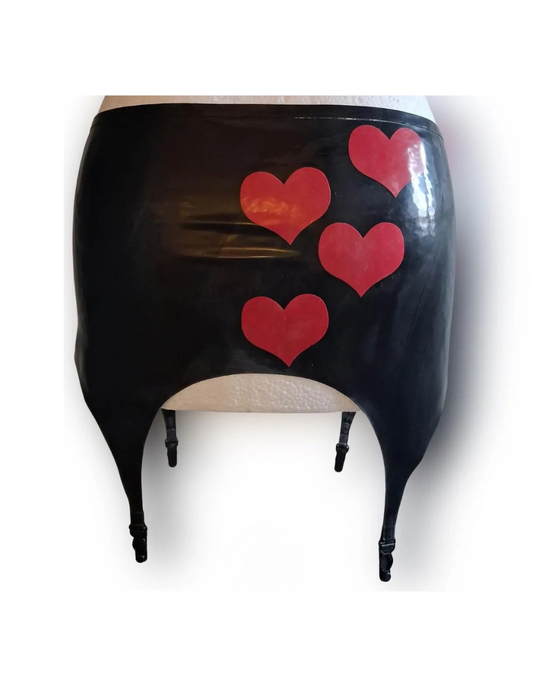 A black latex suspender belt with four red heart designs on the front, displayed on a white mannequin torso against a white background.